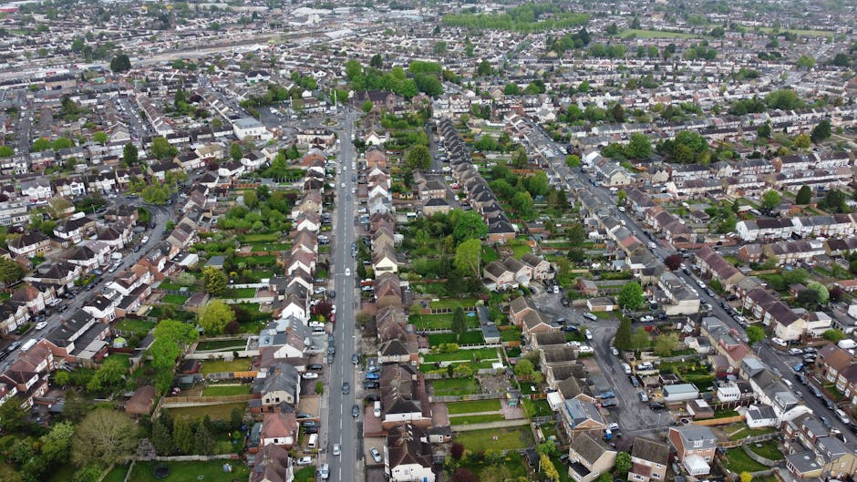An aerial view of a suburban residential area showing a grid of streets lined with terraced and semi-detached houses with varied roof styles, gardens, and driveways. The gardens feature a mix of lush 
